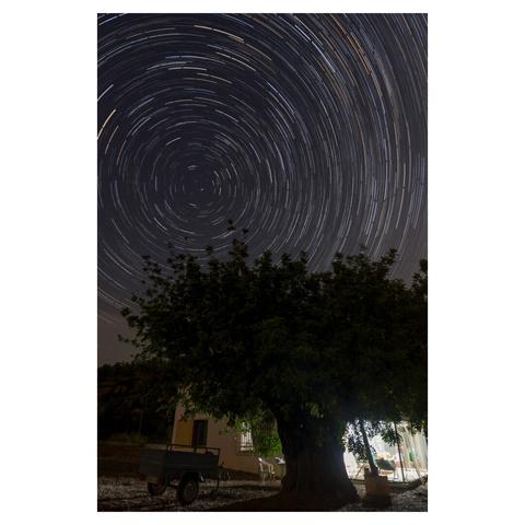 Foto nocturna circumpolar de estrellas sobre una casa rústica de campo con el porche iluminado y una garrofera centenaria. Cerca del centro cruza una estrella fugaz muy tenue.