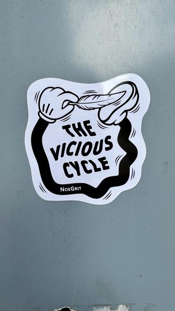 Sticker on the back of a road sign representing a cartoonish hand linked to foot by a black limb and is ticking the foot with a feather. The text on the sticker says «  The Vicious Cycle » by NoeGrit