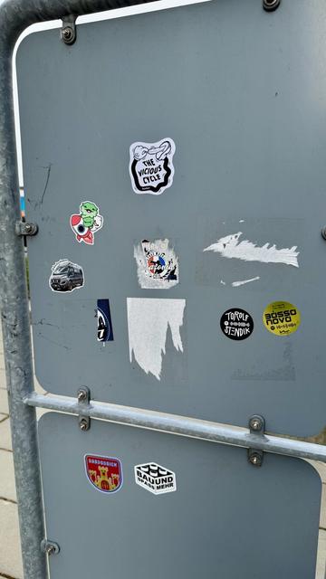 Sticker on the back of a road sign representing a cartoonish hand linked to foot by a black limb and is ticking the foot with a feather. The text on the sticker says «  The Vicious Cycle » by NoeGrit