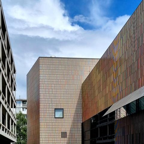 On the left, the concrete façade of a university building, right next to it the multi-colored façade of the Brandhorst Museum building, consisting of ceramic rods that echo the colors of the surrounding buildings. Above, a cloudy sky with a few patches of blue.