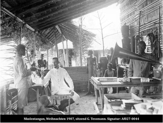 Das Foto von 1907 zeigt den weiß gekleideten Günter Tessmann auf einer überdachten Veranda auf einem Stuhl neben einem Grammophon sitzend. Um ihn herum gruppieren sich mehrere Einheimische.