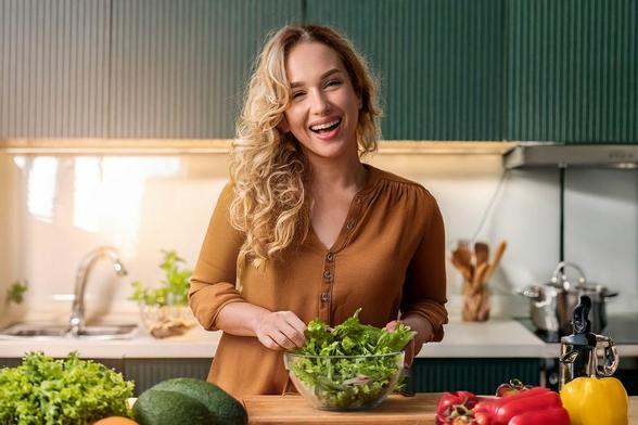 Mujer sonriente preparando una ensalada fresca en una cocina moderna, rodeada de verduras como lechuga, aguacate y pimientos morrones. Aunque sea una simple ensalada, preparar comida despierta la creatividad. Imagen GIA.