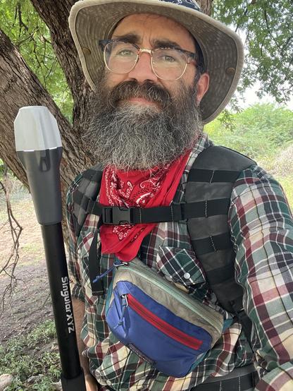 A selfie of a man wearing a hat and bandana and a backpack holding a survey instrument in one hand and with a blue small Patagonia fanny pack strapped to his chest.