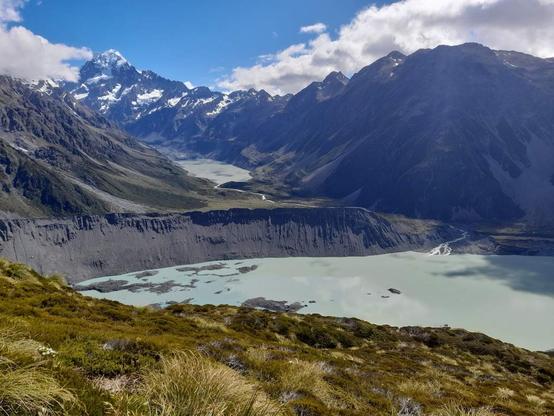 Au centre de cette photo, le mont Cook (Aoraki). Le plus haut sommet de Nouvelle-Zélande, s’élève sous un ciel bleu clair partiellement voilé de nuages. Sa cime enneigée perce les nuages, dominant une chaîne de montagnes aux pentes escarpées, parfois sombres, parfois illuminés par les rayons du soleil.
Au premier plan, des plantes alpines et des touffes d’herbe dorée recouvrent une pente douce. En contrebas, un vaste lac glaciaire – aux eaux couleur turquoise pâle et laiteuses – s’étend paisiblement. Ce lac est bordé par un impressionnant rempart rocheux, formé par une moraine glaciaire, comme une falaise grise qui barre l’accès direct au massif.
En arrière-plan, une vallée glaciaire sinueuse remonte entre les montagnes, où serpente une rivière alimentée par la fonte des glaciers. L’ensemble compose un paysage spectaculaire, sauvage et intact, typique des Alpes du Sud néo-zélandaises.