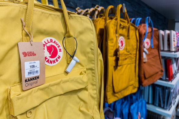 Ein gelber Rucksack der Marke Fjällräven Kånken in einem Ladenregal. Am Griff hängt ein braunes Preisschild mit dem Logo von Fjällräven und der Aufschrift: „KÅNKEN Original – Coop City – 109 Franken“. Im Hintergrund hängen weitere Rucksäcke in Gelb, Braun und Blau.