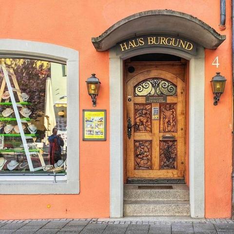 A richly carved wooden door with various scenes and a decorative metal grate in its upper panel is set into an orange building. Above the door, a curved awning reads "HAUS BURGUND." Two vintage-style lamps flank the door, and a shop window is to the left, reflecting a person and displays. The number "4" is visible to the right of the door.