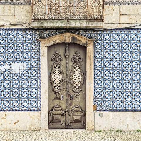 An elaborately carved light brown wooden door with decorative metal accents is set into a stone frame. The wall around the door is covered in blue and white geometric patterned tiles. Above the door, a rusted, ornate metal balcony railing is visible against a plain wall.
