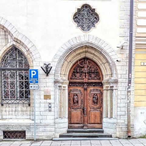 A grand, ornate wooden door with intricate carvings of figures and patterns, set within a tall arched stone doorway. Above it, a decorative iron grate is set into a round window, and another carved window is to the left, all part of a light-colored stone building. A blue parking sign is on the left.
