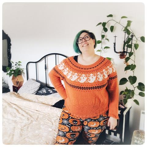 je pose dans ma chambre (on voit le lit avec des draps blanc et jaune, une plante pothos entoure en fond une partie de mon corps, une autre plante est visible en fond), je porte un pull orange tricoté avec des fantômes blanc, j'ai les cheveux vert au carré, de grandes lunettes et un pantalon halloween
