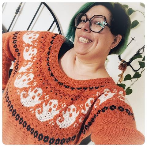 je pose dans ma chambre je porte un pull orange tricoté avec des fantômes blanc, j'ai les cheveux vert au carré, de grandes lunettes et un grand sourire