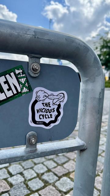 Sticker on the back of a road sign representing a cartoonish hand linked to foot by a black limb and is ticking the foot with a feather. The text on the sticker says «  The Vicious Cycle » by NoeGrit
