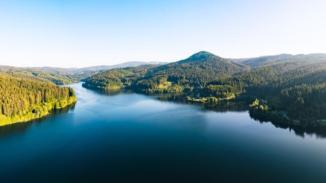 Das Bild zeigt eine beeindruckende Panoramaaufnahme einer Seen- und Waldlandschaft. Die Szene wirkt ruhig, idyllisch und nahezu unberührt.
Im Vordergrund: Ein ruhiger, tiefblauer See erstreckt sich in die Ferne. Das Wasser ist spiegelglatt und reflektiert leicht den Himmel sowie die umliegenden Wälder.
In der Mitte: Dichte, sattgrüne Nadelwälder bedecken die sanft geschwungenen Hügel auf beiden Seiten des Sees. Die Uferlinie ist weitgehend naturbelassen, mit nur wenigen kleinen Gebäuden oder Anzeichen menschlicher Präsenz.
Im Hintergrund: Die Landschaft geht in eine hügelige, teils bergige Region über, wobei ein markanter Berg in der rechten Bildmitte herausragt. Weiter hinten sind noch weitere, sanfte Hügel zu erkennen.
Der Himmel ist klar und wolkenlos – ein leuchtendes Blau, das auf schönes Wetter und gute Sichtbedingungen hinweist. Die Lichtverhältnisse lassen auf frühe Morgenstunden oder späten Nachmittag schließen, da das Licht warm ist und aus schrägem Winkel kommt.