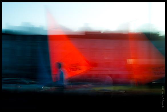 Abstract long exposure photograph showing motion blur of a person walking in front of a bright red boat sail in Kinsale harbour, County Cork, creating streaks of red, blue and white colours.
