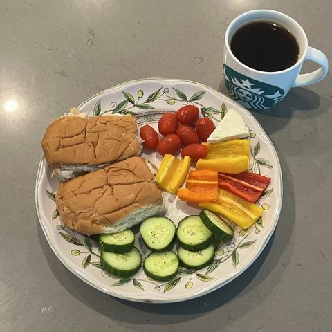 A plate of sliced bell peppers and cucumbers, whole grape tomatoes, a piece of cheese, two brioche buns, and a cup of coffee