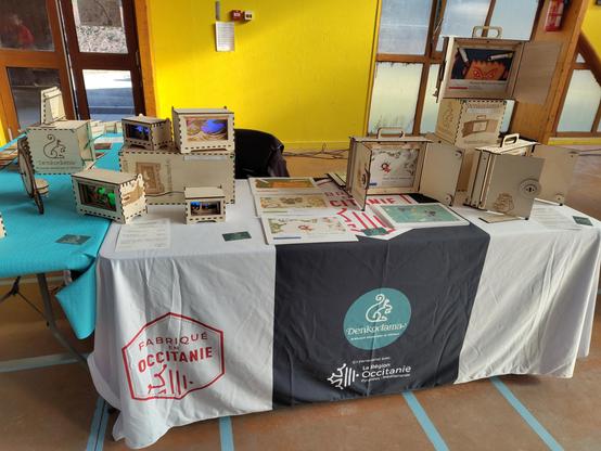 Deux tables couvertes de nappes, pour un stand Denkodama (logo sur fond vert avec un écureuil tracé en blanc visible sur la partie de la nappe qui pend devant la table). Sur les tables, divers objets en bois : trois kamishibai à droite, des décorations lumineuses de type lightbox à gauche. Au fond, un mur jaune