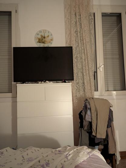 A slightly underexposed, eye-level, full shot of a bedroom at night. A large, dark TV is sitting on top of a tall, white dresser with four large drawers and two small drawers. Above the TV is a round clock with a cat. To the right of the dresser is a pile of clothes draped over a chair. Two tall windows are visible in the background, with the window on the right partially covered by a thin, floral-patterned curtain. The bed in the foreground has a white and purple floral comforter.