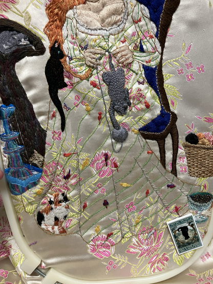 A section of an embroidered picture showing a woman in a long gown decorated with vines, birds, and butterflies, knitting a cat while surrounded by more cats.