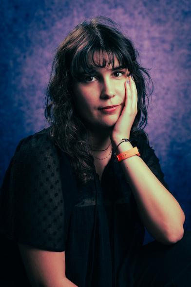 A person with wavy hair resting their head on their hand is wearing an orange watch and a black outfit. The background is a mottled blue and purple, enhancing the contemplative expression.