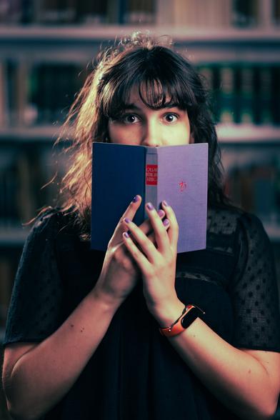 A person with long, wavy hair stands in a library, holding an open book close to their face, with eyes wide in a look of surprise or intrigue. They wear a black shirt and an orange watch. Bookshelves filled with books line the background.