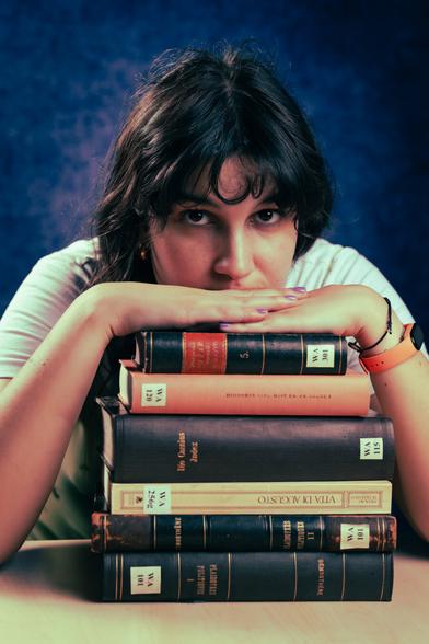A person rests their chin on a stack of thick, vintage books, gazing intently at the camera. They wear a white shirt and have an orange watch on their wrist. The background is a textured deep blue, contrasting with the books’ earthy tones.