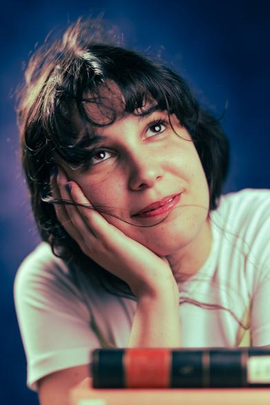 A person with short, dark hair and a thoughtful expression gazes upward. They are resting their face on their hands, with a soft smile. The background is a deep blue, and a stack of books is in the foreground.