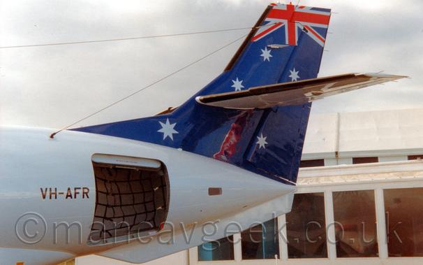 Side view of the rear fuselage and tail of a twin propellor-engined airliner parked facing to the right with the cargo hold door open.
The plane is mostly white, with a dark blue tail with 5 white stars scattered around, and part of a British flag at the top.
The black registration "VH-AFR" is in front of the open cargo door.
In the background, a heavily glazed, low white building is on the lower right of the frame, with flat white sky fills the rest of the frame.