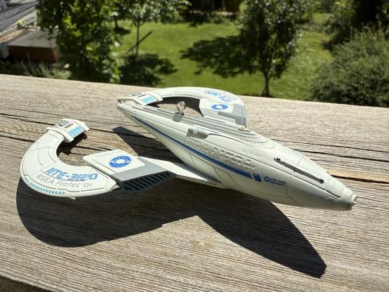 A model of a futuristic spaceship, featuring a sleek design in light gray with blue accents. The ship is positioned on a wooden surface, with greenery in the background, suggesting an outdoor setting.