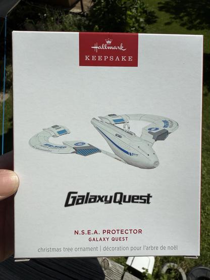 The image features a Hallmark Keepsake box containing a Christmas tree ornament modeled after the N.S.E.A. Protector spaceship from the movie "Galaxy Quest." The box is predominantly white with colorful graphics of the spaceship and includes both English and French text about
