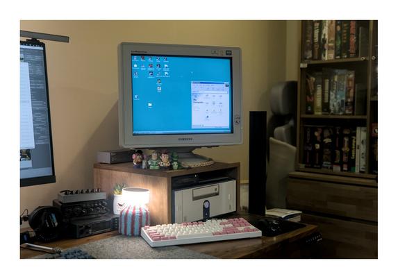 Photo of the right-hand side of my desk where my Pentium 3 Windows 98 PC stands. It's a small case that can be used in both tower and desktop configuration that is laying in the latter configuration underneath a wooden up-side-down-letter-U-shaped wooden stand, on top of which a 19 inch Samsung "SyncMaster" CRT monitor stands. To the left of the PC one can find a couple of audio related components, including a 6-input basic mixer, an XLR USB Audio interface, and a Roland SoundCanvas SC-88vl. My Sony MDR-7506 headphones can be seen slightly to the left of this audio stack. To the left and right behind the monitor a small metal box can be seen, which is an active VGA duplicator. In front of the CRT are several small Toy Story toy characters. Underneath the wooden stand and right on top of the PC case an Iomega ZIP 100 drive can be seen. In front of the PC are a  black PS2 mouse that feels all kinds of cheap, and one of my mechanical keyboards (a Leopold FC750R in white and light pink). In the background towards the right a cabinet can be seen, showing a handful of big box DOS and Windows games.