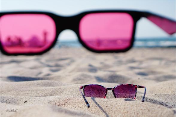 Im Sand am Strand liegt im Vordergrund im Fokus eine Sonnenbrille mit rosa getönten Gläsern. Ein paar Meter weiter liegt eine menschengroße Sonnenbrille im Sand, ebenfalls mit rosa getönten Gläsern. Durch ihr linkes Glas zeichnen sich die Umrisse einer Gruppe Menschen ab. Im Hintergrund ist das Meer zu erkennen.