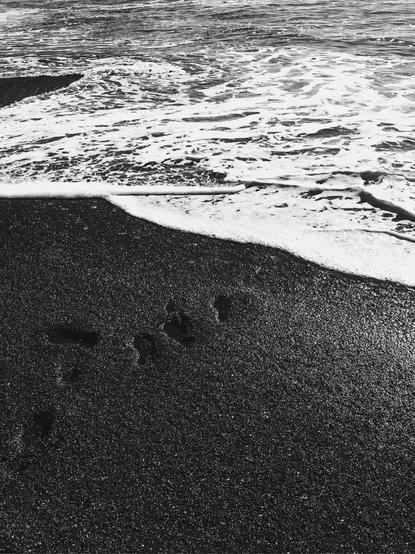 A black and white photo of footprints in sand coming from the bottom left corner leading diagonally towards the opposite corner and meeting an incoming wave of white foamy ocean water