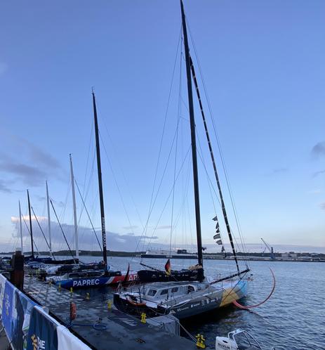 Sechs Imoca-Segelboote liegen an einem Ponton zum Start bereit. Im Hintergrund noch eine Fähre.