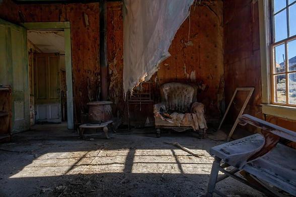 An eerie abandoned living room in Bodie, California's ghost town. Sunlight streams through broken windows, revealing decaying furniture and relics from a time long gone.