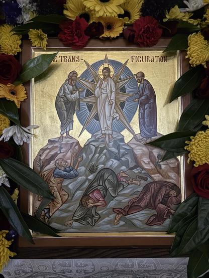 An icon of the Transfiguration of Christ, surrounded by freshly-cut flowers. Christ is shown in glory with Moses and Elijah, while Peter, James, and John fall down in amazement.
