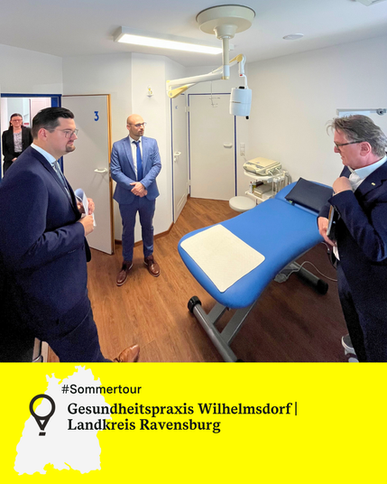 Eine Frau und drei Männer in Anzügen, darunter Minister Manne Lucha, stehen in einem Behandlungszimmer mit blauer Liege und medizinischem Gerät an der Decke. Text: Gesundheitspraxis Wilhelmsdorf, Landkreis Ravensburg.