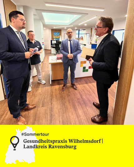 Vier Männer stehen am Empfang einer Arztpraxis und unterhalten sich, darunter Minister Manne Lucha. Text: Gesundheitspraxis Wilhelmsdorf, Landkreis Ravensburg.