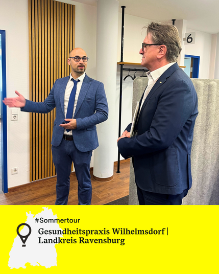 Zwei Männer stehen nebeneinander in einem Raum, einer davon ist Minister Manne Lucha. Der andere Mann erklärt etwas und zeigt etwas mit seinen Händen. Text: Gesundheitspraxis Wilhelmsdorf, Landkreis Ravensburg.