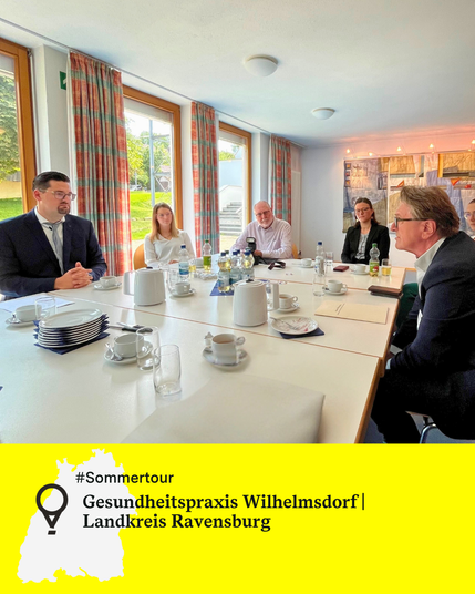 Sechs Personen sitzen an einem Tisch und unterhalten sich, auf dem Tisch stehen Kaffeetassen und Kannen. Rechts im Bild ist Minister Manne Lucha, er beugt sich vor und hört zu. Text: Gesundheitspraxis Wilhelmsdorf, Landkreis Ravensburg.