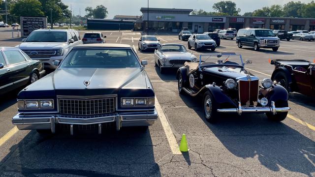 Photograph in a parking lot of a dark blue mid-70s Cadillac Fleetwood Brougham seen from the front next to a small maroon 1952 MG TD to the right.