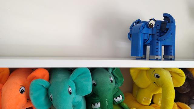 A elephpant made out of Lego stands atop a white shelf. A bunch of colourful elephpants are on the shelf beneath it.