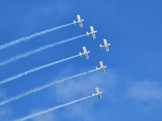 A dynamic shot of six small white planes flying in diagonal formation across a clear blue sky. Each plane is leaving a long, distinct white smoke trail behind it, creating parallel lines that fan out across the frame.