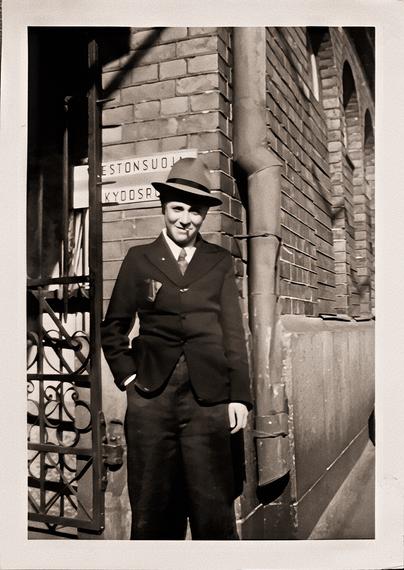Black and white slightly sepia-toned picture of a teenage boy dressed in a suit and hat. He's got one hand in his pocket and what looks like a cigarette in his mouth, smiling a sort of a wry smile.

He's standing in front of a brick building and there's a wrought iron gate next to him, and behind his head there's a partially obscured bilingual sign that reads "[vä]estönsuoj[a]" in Finnish, "[s]kyddsr[um]" in Swedish (obscured letters marked with brackets)