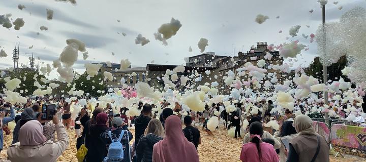 A view of Centenary Square with different sized blobs of coloured foam (soap and water with food colouring) filling the sir abovr. Below, crowds of people watch on some with cameras filming.