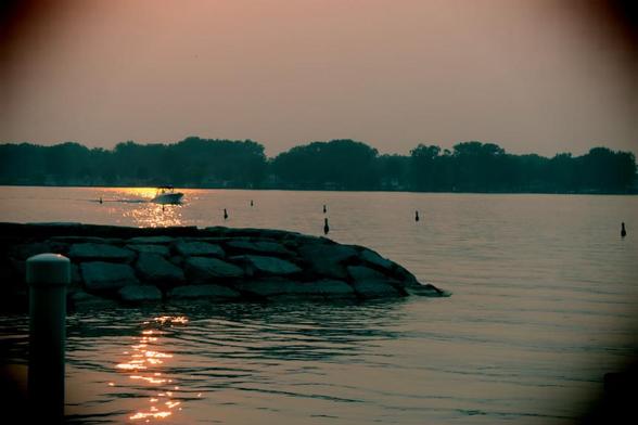 Looking out across the St Clair River from the Canadian side, viewing a recreational boat returning through channel markers to the dock, as the setting sun's light ripples in the water.  The wooded shoreline on the American side shows nothing of the country's decline into authoritarianism and Fascism.