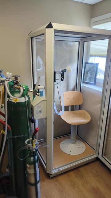 An unoccupied pulmonary function test machine. The machine itself is a large metal and glass box, with a seat and mouthpiece inside. Large tanks of air and oxygen are connected to the machine.
