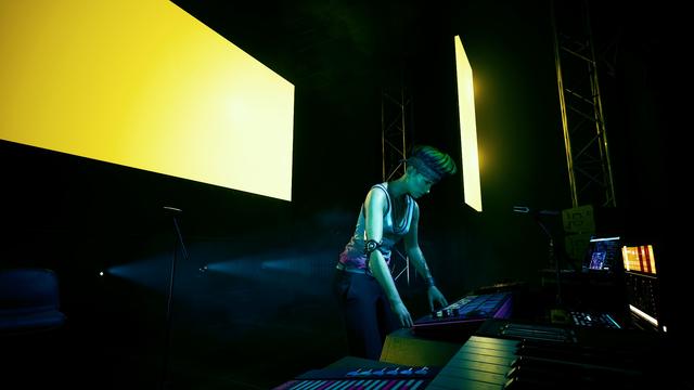 Une Dj aux platines avec un écran jaune derrière.
Au tout premier plan, on peu apercevoir un clavier (de piano)