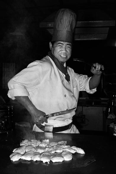 A chef in a white uniform skillfully prepares food on a grill, surrounded by steam, with a knife in hand and freshly cooked items in front.