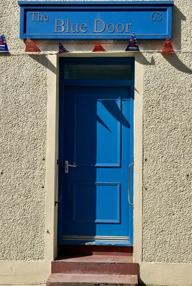 A bright blue door with a decorative sign above in the same shade of blue reading "The Blue Door 63." The door is framed by a textured wall and has a small set of maroon steps leading up to it. Colorful triangular bunting is strung above the door.