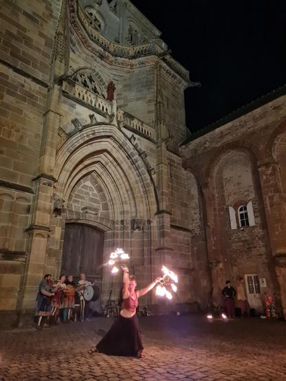Sur le parvis d'une église prieurale, une femme fait un spectacle de feu tandis que des musiciens jouent en arrière-plan.