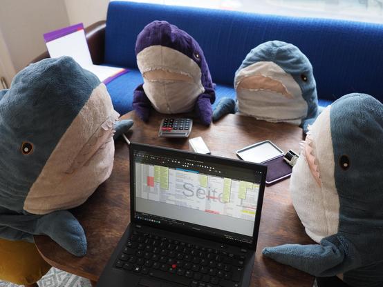 Vier Plüschtier-Haie sitzen um einen Tisch. Auf diesem liegen ein Stempel mit Stempelkissen, ein Taschenrechner und ein Laptop, auf dessen Bildschirm ein Tabellendokument angezeigt wird.